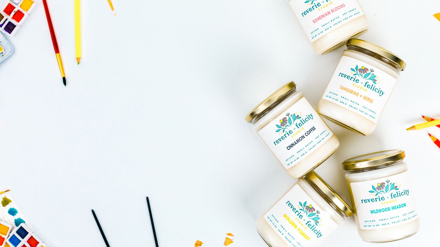 Reverie + Felicity Studio soy wax candles in a flatlay with sharpened colored pencils, paintbrushes and watercolor paints on a white background