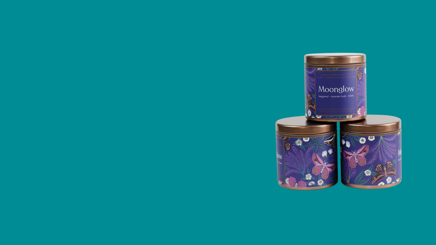 Three Moonglow soy wax candles in lidded bronze tins with purple labels featuring an illustration of moths, lavender springs, greenery and whilte flowers are stacked in a triangle on a teal background